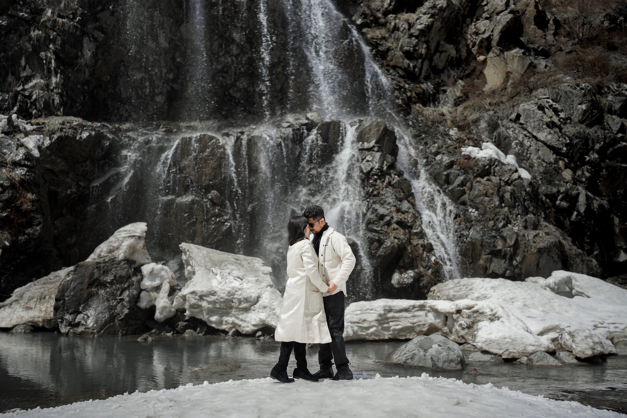 A couple embracing the winter scenery, touching foreheads as a waterfall creates a soothing ambiance in the background.